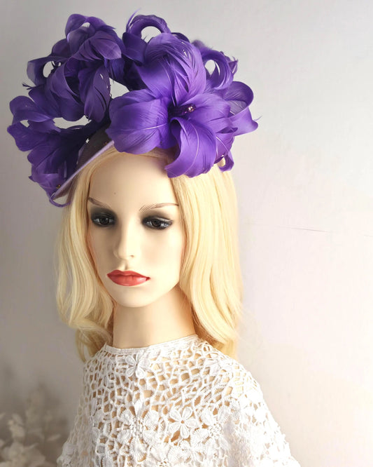 A large purple fascinator hat with layered artificial flowers, displayed on a blonde mannequin head wearing a white lace top against a plain light background.
