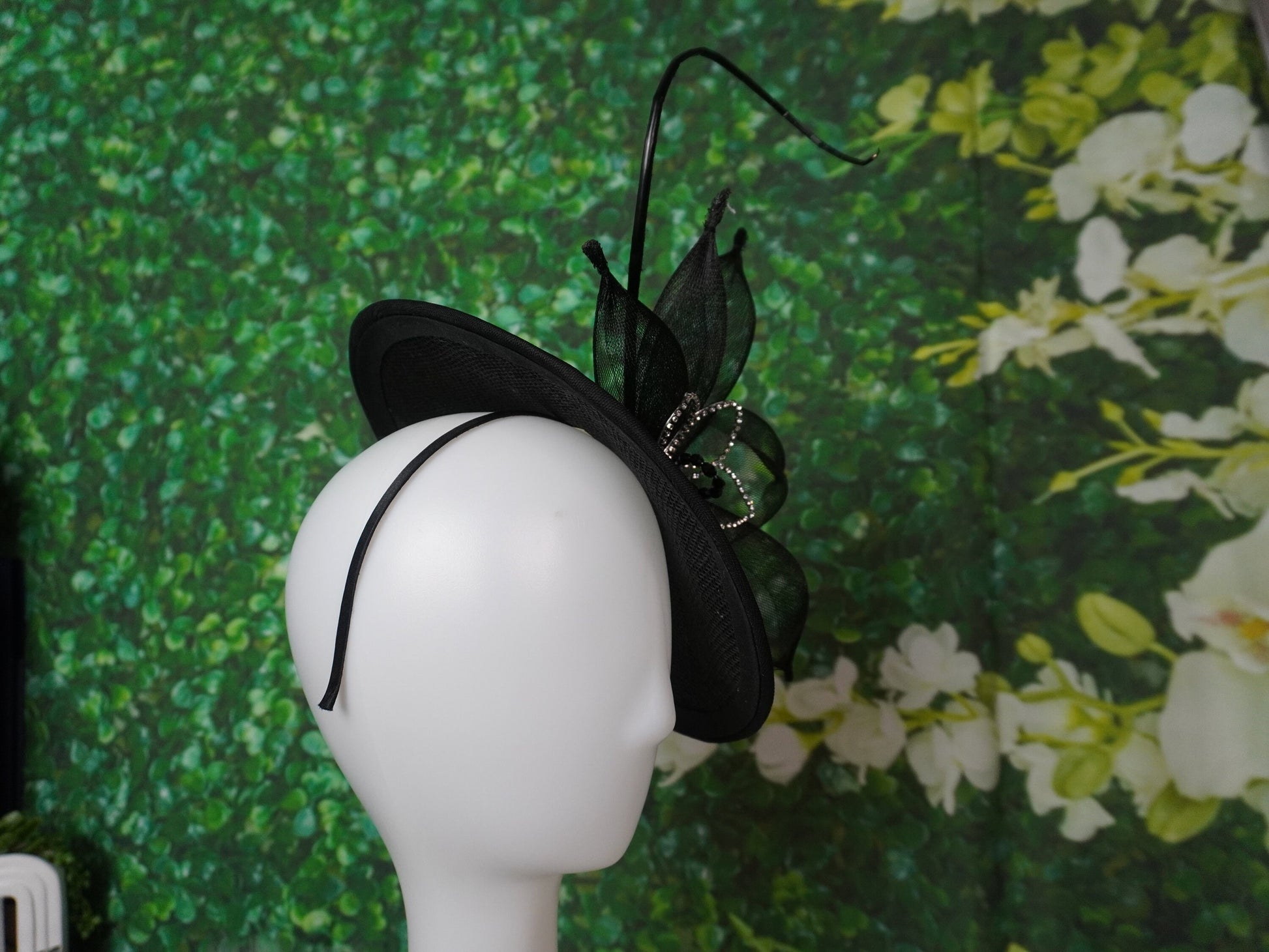 A black fascinator hat with sheer netting and a curved feather accent, displayed on a white mannequin head against a green leafy floral background.