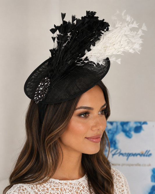 A woman wearing a black fascinator hat with black and white feathers and a jeweled embellishment, photographed against a light neutral background.