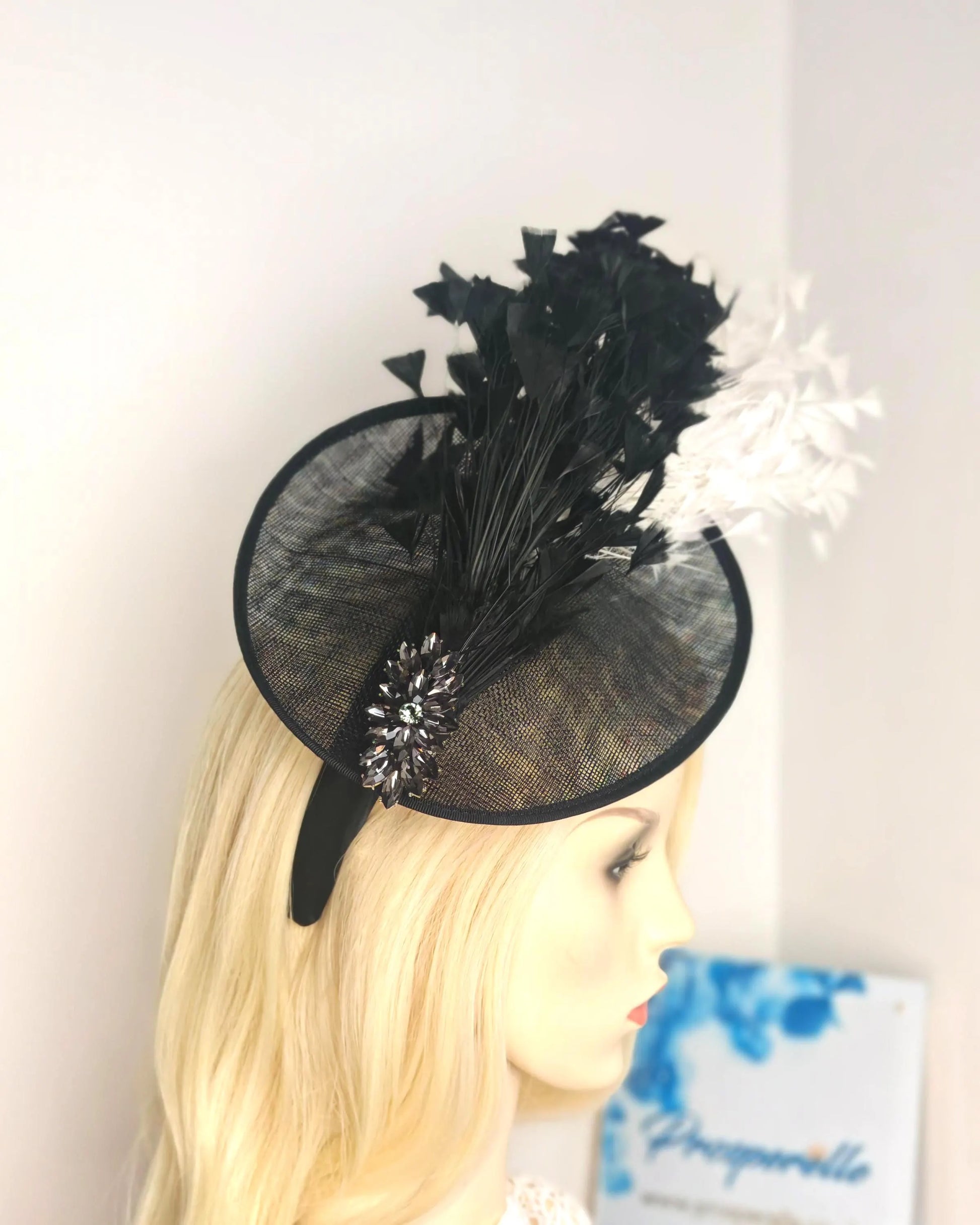 A black fascinator hat with black mesh, black feathers, and a white feather accent, displayed on a blonde mannequin head against a plain white wall.