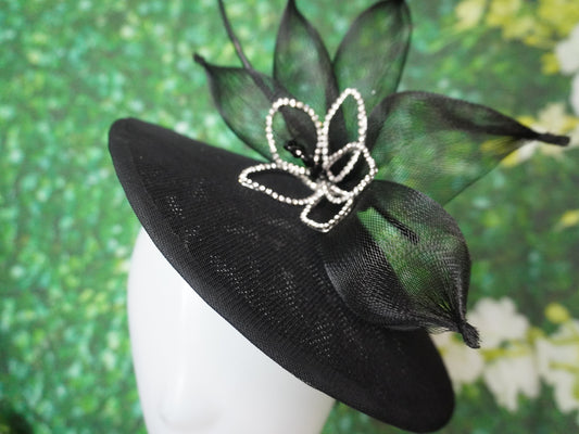 A black fascinator hat with a wide brim, decorated with black mesh leaves and a silver rhinestone-embellished bow, displayed on a white mannequin head against a green textured background.