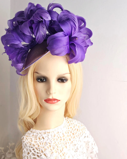 A large purple fascinator hat with layered fabric flowers and a dark brim, displayed on a blonde mannequin head against a plain light background.