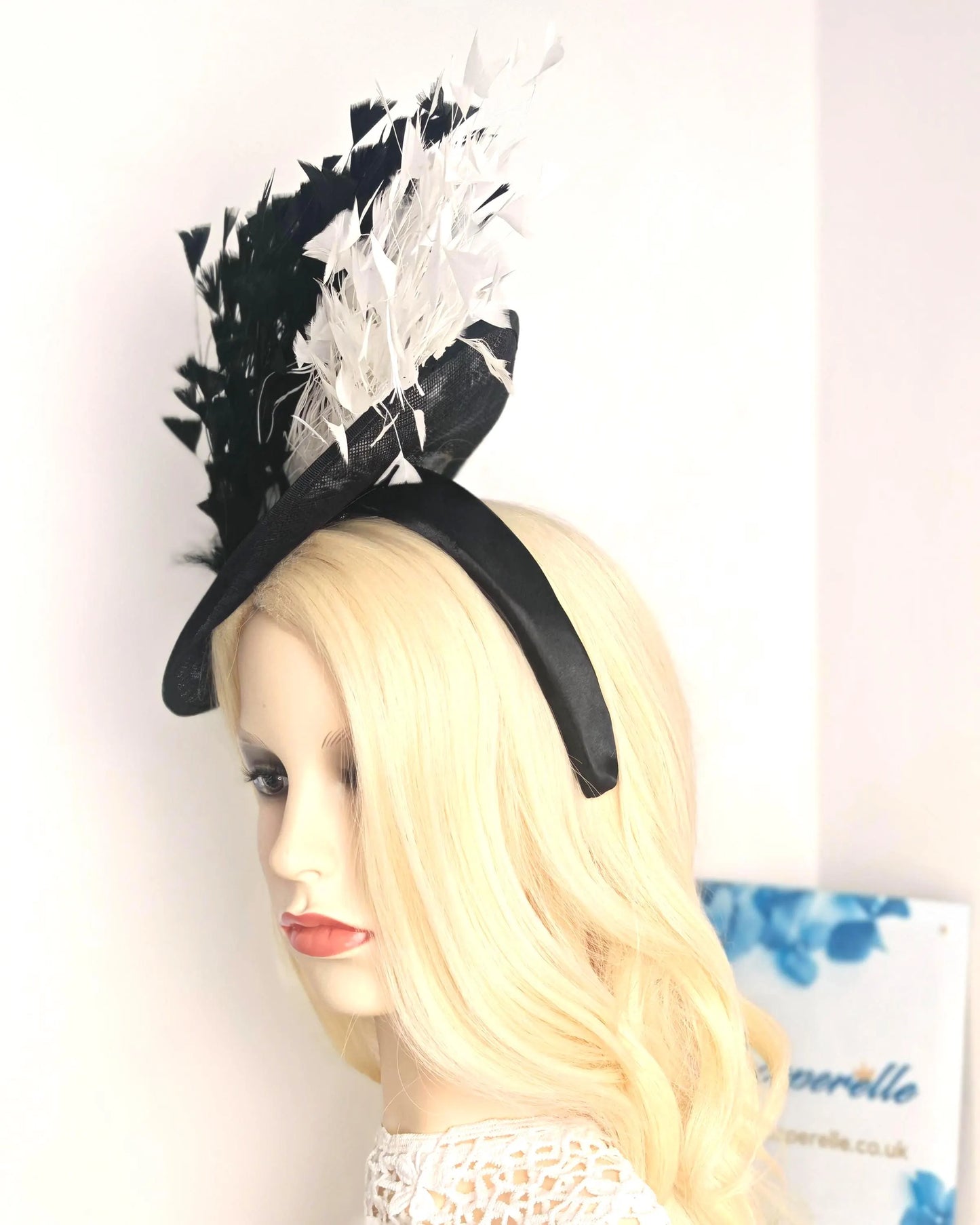 A black fascinator hat with white feather accents, displayed on a blonde mannequin head against a plain white wall.